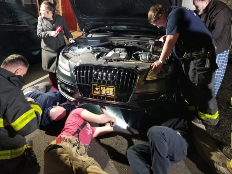 Firefighters under and above the hood of a car.